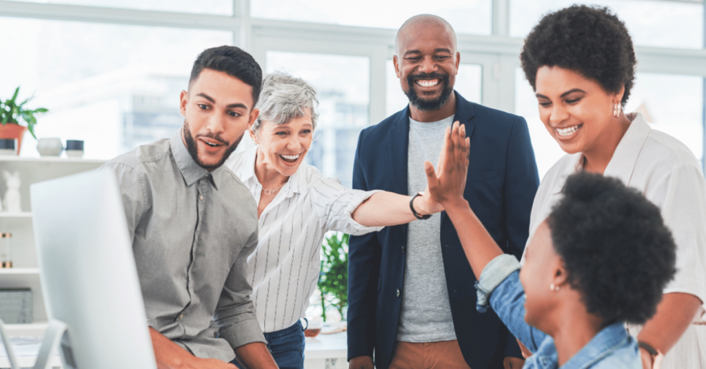 Photo of five coworkers smiling at each other around a computer. Two are giving each other a high five.