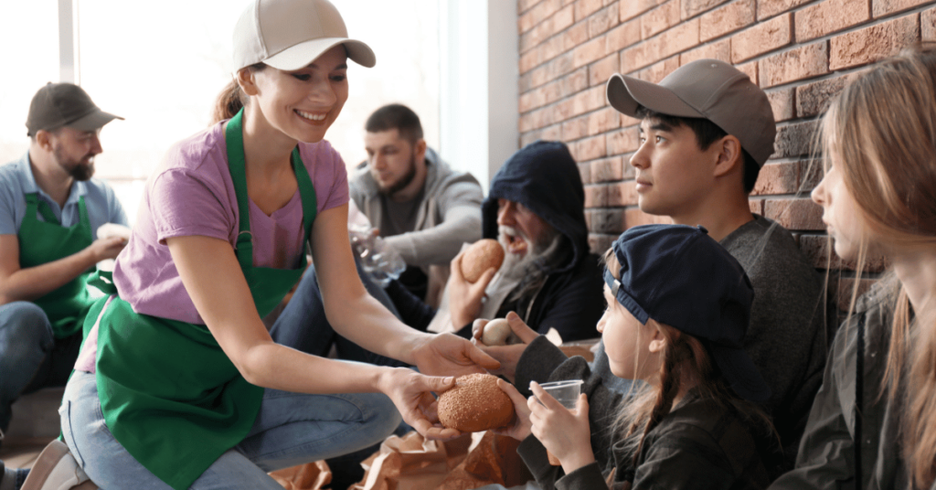 Photo of a volunteer smiling while handing out food to people sitting against a brick wall