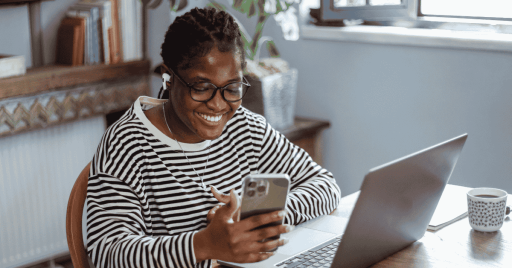 Smiling remote worker wearing headphones and glasses while looking at her phone at a home office desk.